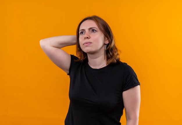 Nadenkende jonge toevallige vrouw die hand achter hoofd op geïsoleerde oranje muur zet