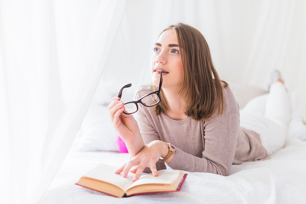 Gratis foto nadenkende jonge vrouw die zwarte oogglazen in haar mond bijt die op bed ligt