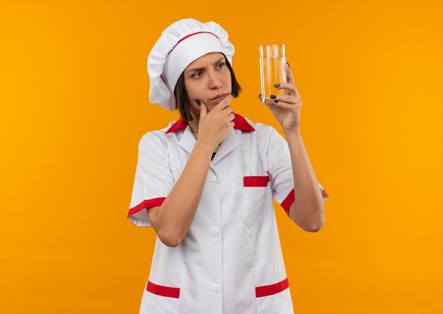 Nadenkende jonge vrouwelijke kok in chef-kok uniform houden en kijken naar glas water met hand op kin geïsoleerd op oranje muur