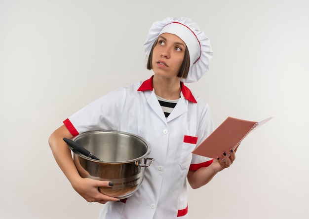 Nadenkende jonge vrouwelijke kok in de pot en het blocnote van de chef-kok de eenvormige holding die omhoog geïsoleerd op witte muur kijken