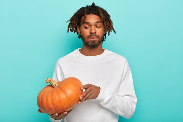 Nadenkende man met dreadlocks, houdt pompoen vast, bereidt zich voor op Halloween, draagt een witte trui, heeft een baard