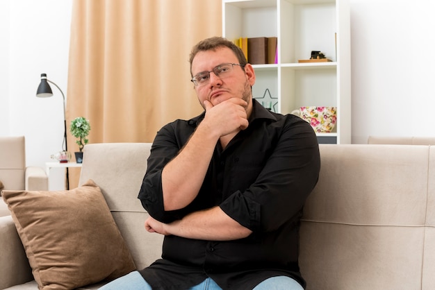 Nadenkende volwassen Slavische man in optische bril zit op fauteuil kin vast te houden en kant in de woonkamer te kijken