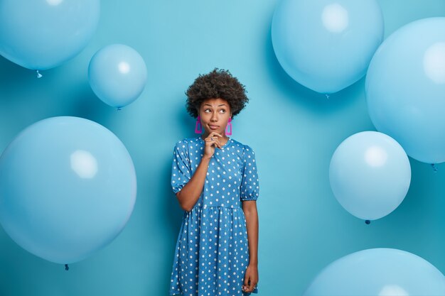 Nadenkende vrouw met donkere huid draagt blauwe polka dot jurk, op ballonfeest, gekleed in modieuze outfit, heeft een peinzende uitdrukking, gaat haar verjaardag vieren. Vrije tijd, feesten concept