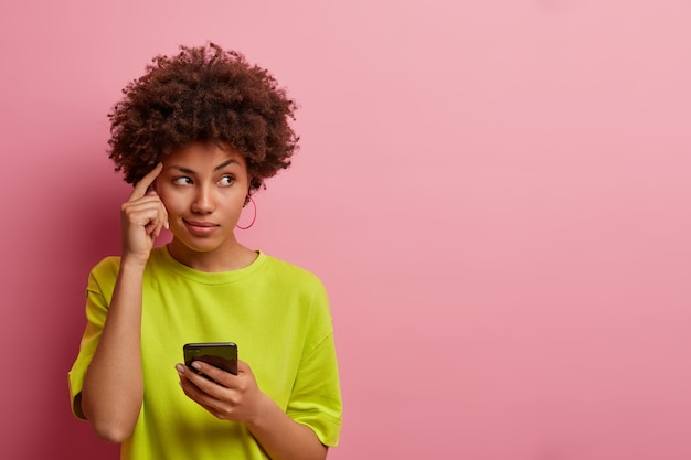 Nadenkende vrouw met donkere huid houdt vinger op slaap, kijkt bedachtzaam opzij, gebruikt nieuwe gadget voor de eerste keer, kleedt zich nonchalant aan, probeert de naam van het product te onthouden voordat ze op internet surft, heeft een idee