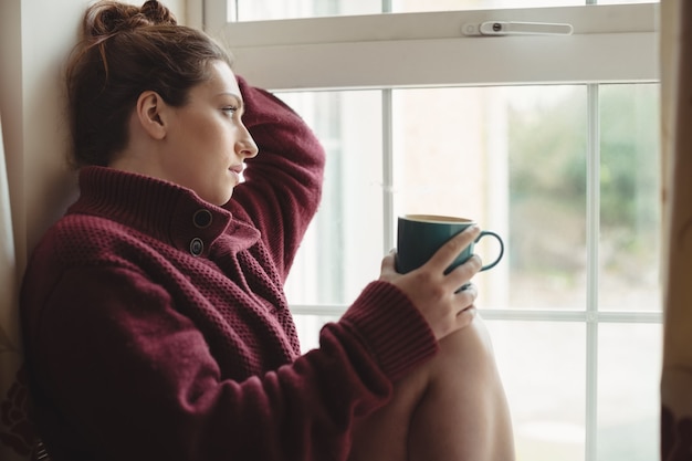 Nadenkende vrouw zit op de vensterbank en koffiekopje te houden