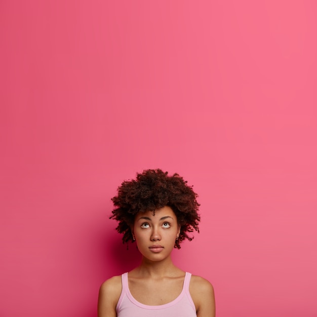 Gratis foto natuurlijk gekrulde afro-amerikaanse vrouw kijkt bedachtzaam naar boven, heeft wat in gedachten, draagt vrijetijdskleding, staat tegen een roze muur, kopieert ruimte voor uw promotie, herinnert zich iets