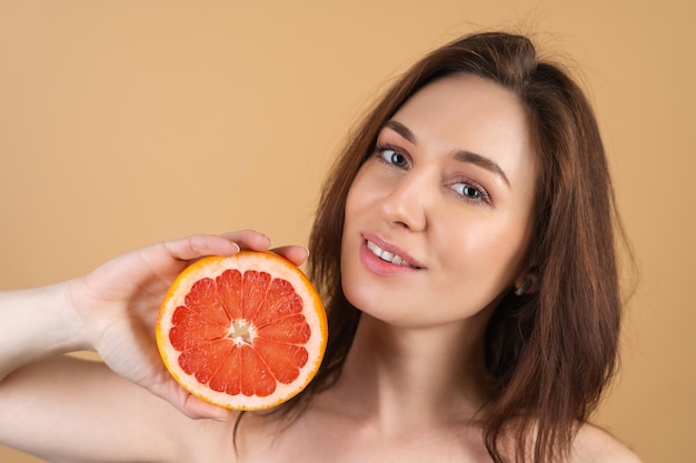 Nauw schoonheidsportret van een topless vrouw met perfecte huid en natuurlijke make-up, glanzende lippen, op een beige achtergrond met grapefruit