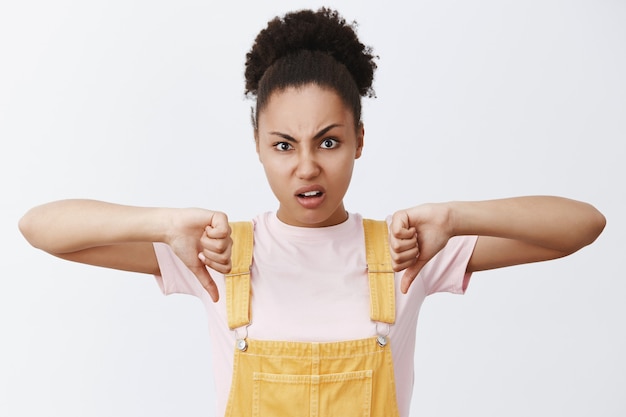 Gratis foto nee, het is een afkeer. portret van ontevreden en teleurgesteld schattige afro-amerikaanse vrouw in trendy gele overall, duimen naar beneden en fronsen van walging