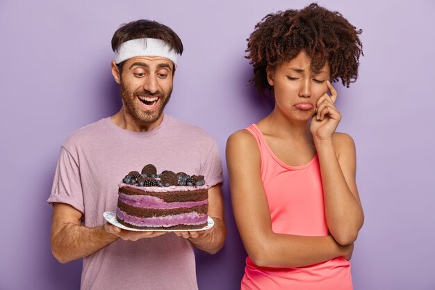 Neerslachtige, boze vrouw verandert van echtgenoot die smakelijke cake op bord houdt, heeft een droevige uitdrukking omdat ze geen zoete desserts kan eten om fit te blijven en slank leidt een gezonde levensstijl, weigert junkfood te eten