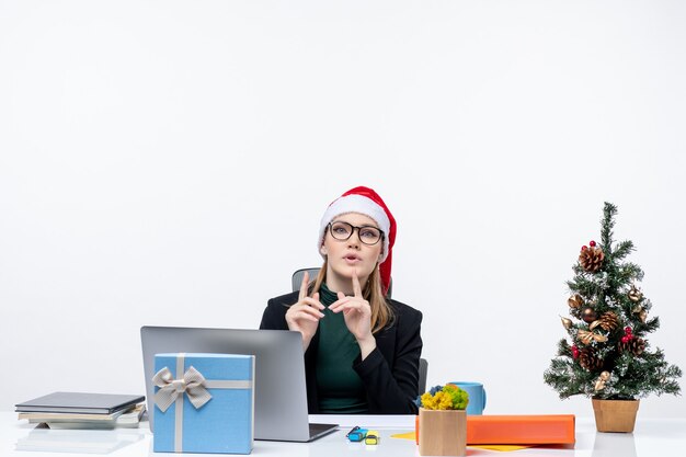 Nerveus zakenvrouw met kerstman hoed zittend aan een tafel met een kerstboom en een cadeau erop in het kantoor op witte achtergrond