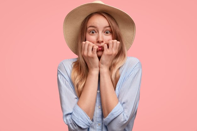 Neurotische mooie dame in hoed en shirt, bijt nagels, staart met angst, voelt zich bezorgd over iets, geïsoleerd over roze muur. Elegante jonge vrouw kijkt zenuwachtig. Mensen en gevoelens