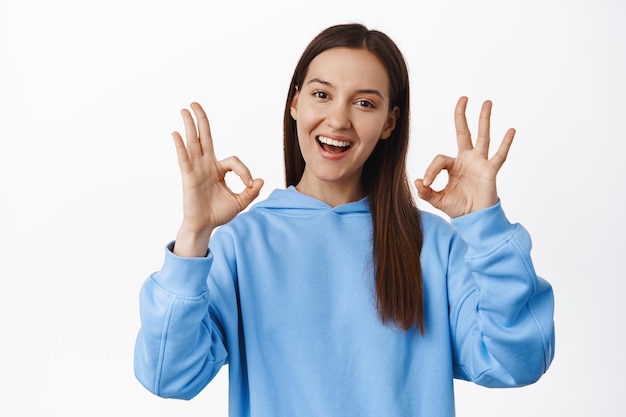 Niet slecht, gefeliciteerd. Glimlachende brunette 20s-vrouw, ok-tekens tonend en tevreden kijkend, prijs geweldige keuze, goed werk, geef goedkeuring, complimenteer, staande op witte achtergrond.