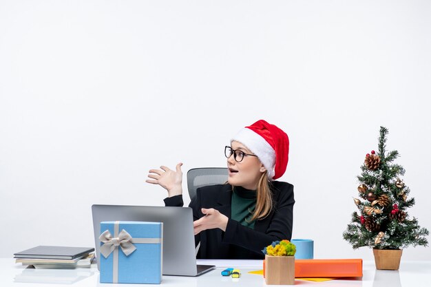 Nieuwjaarsstemming met jonge aantrekkelijke vrouw met een kerstman hoed, zittend aan een tafel met een kerstboom en een cadeau erop en kijken naar iets op kantoor