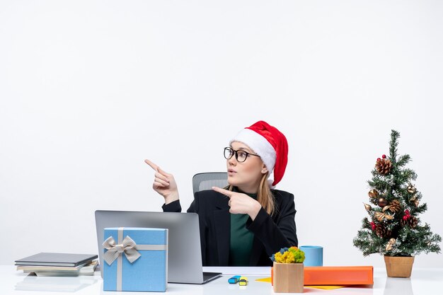 Nieuwjaarsstemming met jonge aantrekkelijke vrouw met een kerstman hoed zittend aan een tafel met een kerstboom en een cadeau erop gericht op iets in het kantoor