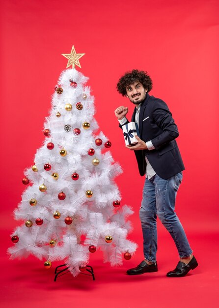 Nieuwjaarsviering met jonge man die zijn geschenk gelukkig toont in de buurt van versierde witte kerstboom op rode beelden