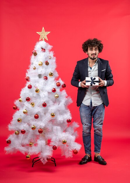 Nieuwjaarsviering met jonge man met een geschenk in de buurt van versierde witte kerstboom op rode beelden