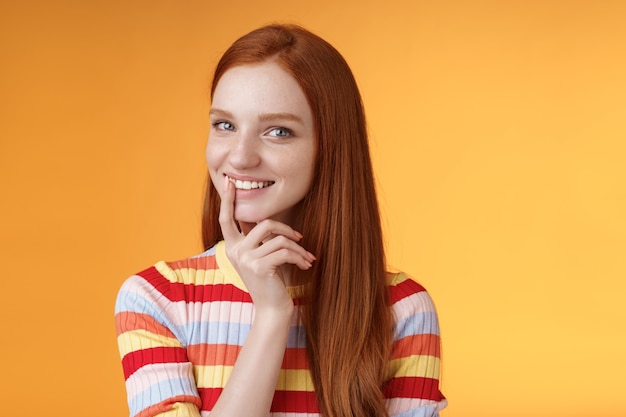 Nieuwsgierig slinkse roodharige jonge 20s vriendin heeft een uitstekend idee grijnzende lastige aanraking lip flirterig mysterieus kijkende camera hebben plannen voor het voorbereiden van interessante verrassing, staande oranje achtergrond.