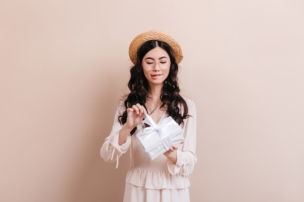 Nieuwsgierige Aziatische vrouw verjaardagscadeau openen. De leuke Chinese gift van de vrouwelijke modelholding op beige achtergrond.