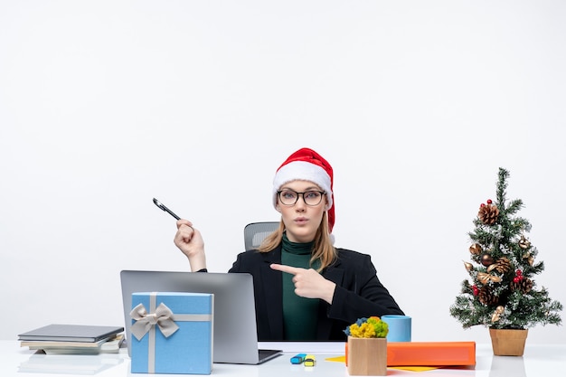 Nieuwsgierige blonde vrouw met een kerstman hoed zittend aan een tafel met een kerstboom en een cadeau erop en iets op witte achtergrond pinting