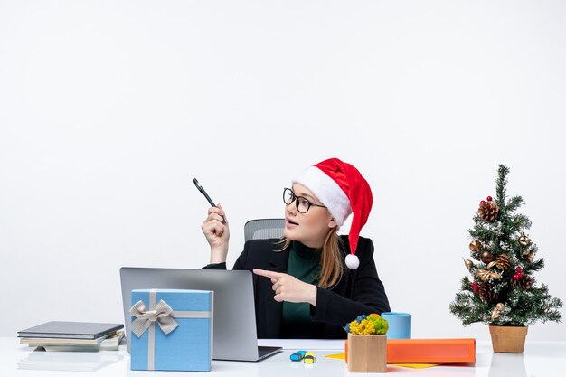 Nieuwsgierige blonde vrouw met een kerstman hoed zittend aan een tafel met een kerstboom en een cadeau erop te kijken naar iets aan de linkerkant op witte achtergrond