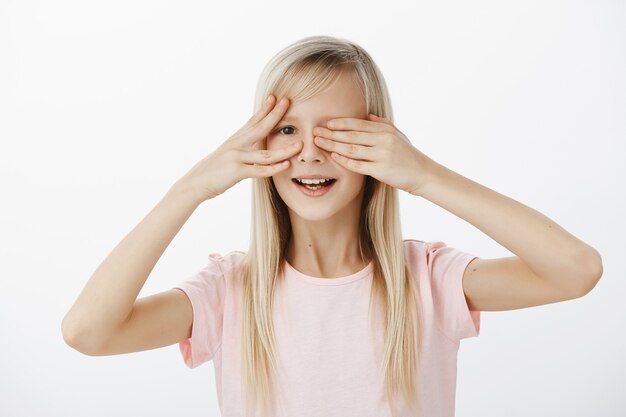 Nieuwsgierige dochter wil zien wat vader voor zijn verjaardag heeft voorbereid. Portret van geïntrigeerd ongeduldig schattig meisje met prachtig blond haar, ogen bedekt met handpalmen en gluren met brede glimlach over grijze muur