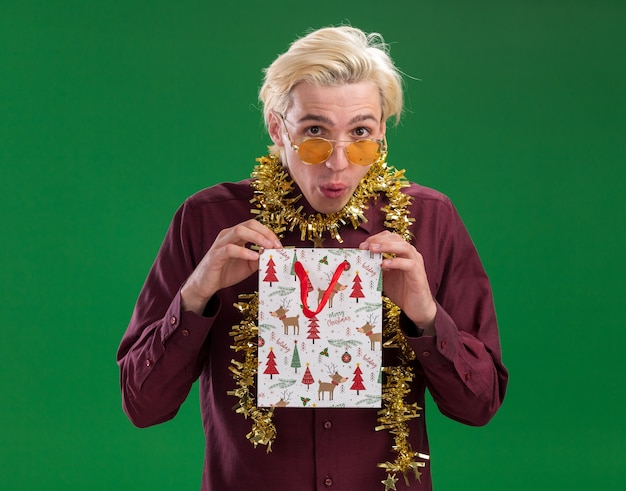 Nieuwsgierige jonge blonde man met bril met klatergoud slinger rond de nek met kerst cadeau zak geïsoleerd op groene muur