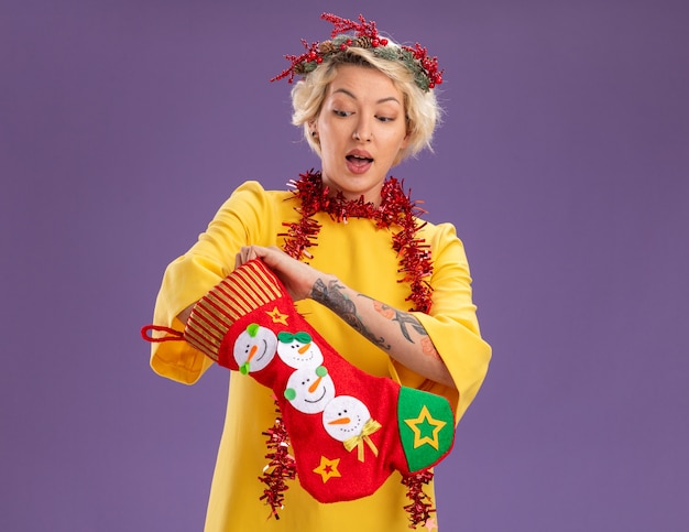 Nieuwsgierige jonge blonde vrouw dragen kerst hoofd krans en klatergoud slinger rond nek houden kerstsok zetten hand erin kijken geïsoleerd op paarse achtergrond