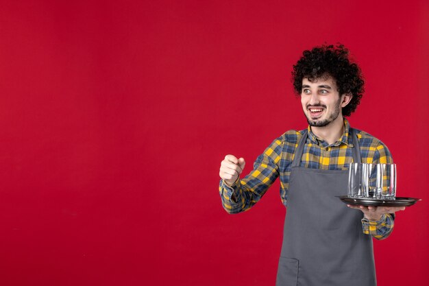 nieuwsgierige ober met krullend haar die een dienblad vasthoudt met een bril gericht op iets op een geïsoleerde rode achtergrond