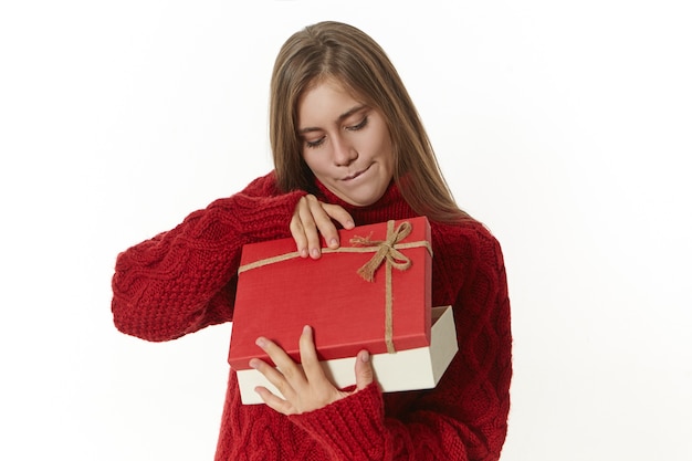 Nieuwsgierige vrouw in gebreide warme trui met mooie geschenkdoos, die ongeduldig opent en met belangstelling naar binnen kijkt.