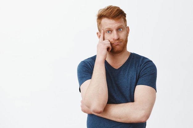 Noodlijdend moe en beu knappe roodharige volwassen man met borstelharen, ademt uit, houdt vinger op de slaap en kijkt neer met een geërgerde en onrustige uitdrukking