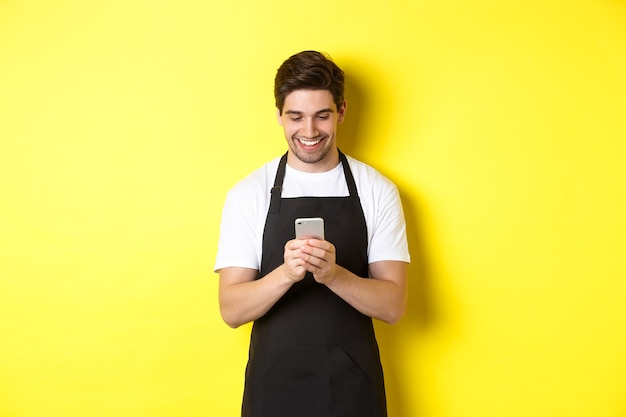 Ober in zwarte schort leest bericht op mobiele telefoon, glimlachend gelukkig, staande over gele achtergrond