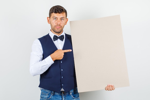 Ober wijzend op leeg canvas in overhemd, vest, spijkerbroek en op zoek naar serieus. vooraanzicht.