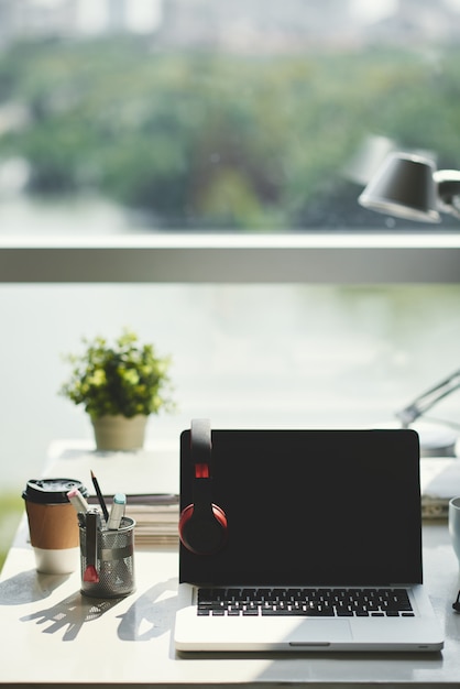 Object shot van uitgeschakelde laptop met open deksel, zwart scherm en koptelefoon, op bureau in kantoor voor raam overdag