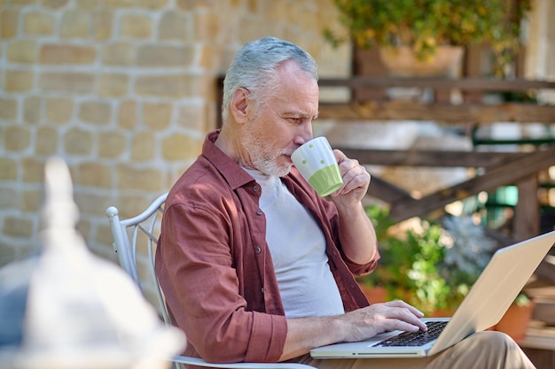 Ochtend thuis. Grijsharige man zit in een fauteuil en leest iets online