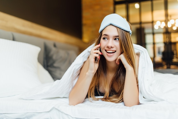 Ochtendbedtijd thuis, blond meisje gebruikt telefoon in blauw slaapmasker