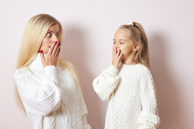Oeps, Omg. Schattig klein meisje en haar jonge moeder beide in witte truien poseren geïsoleerd handen op de mond houden, verbaasd zijn over grote verkoopprijzen, gaan winkelen om kerstcadeautjes te kopen