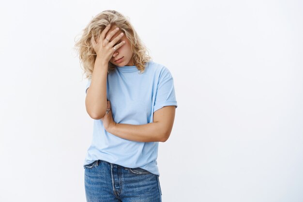 Oh god wat een vernedering. Portret van een beu, uitgeputte en bedroefde jonge vrouw die haar handpalm op het gezicht houdt met teleurstelling, ongelukkig poserend over een witte muur