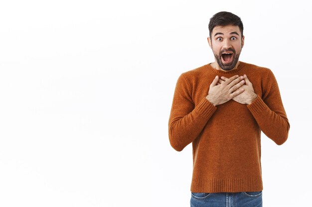 Oh mijn god wat een verrassing. Portret van verbaasde en verbaasde gelukkige bebaarde man houdt handen op het hart, hijgend van opwinding en verbazing, hoor geweldig nieuws, witte muur