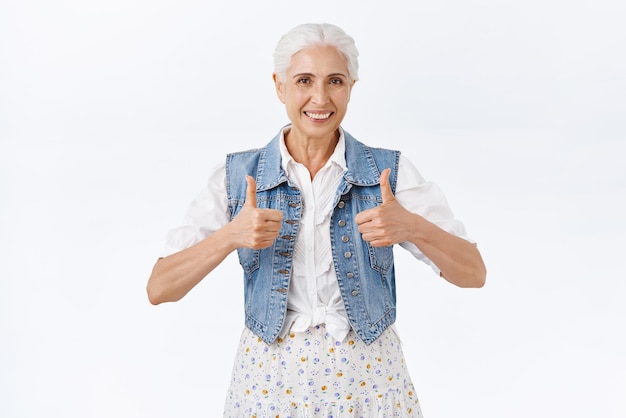 Oma ondersteunt uw goede idee Blij zelfverzekerd lachende gelukkige senior vrouw met gekamd grijs haar in denim vest jurk duim omhoog tonen in goedkeuring accepteren of beoordelen van uitstekende keuze