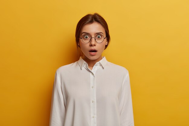Omg, ik geloof het niet! Geschokte emotionele vrouw draagt een grote optische bril en een wit overhemd, reageert op verrassend nieuws, heeft wijd geopende ogen, geïsoleerd over gele muur. Mensen en emoties concept