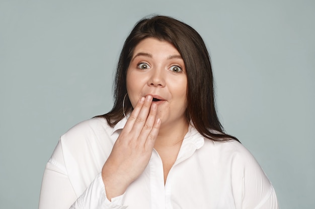 OMG. Menselijke reactie en emoties. Portret van verbaasde, gefascineerde jonge zwaarlijvige blanke vrouwelijke werknemer met mollige wangen voor de mond, geschokt door onverwachte roddels over haar collega