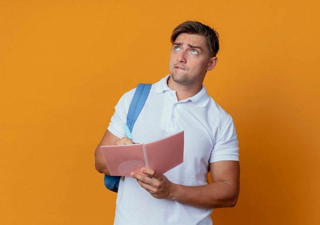 Omhoog kijken verwarde jonge knappe mannelijke student die rugtas draagt die notitieboekje en pen houdt die op oranje achtergrond wordt geïsoleerd