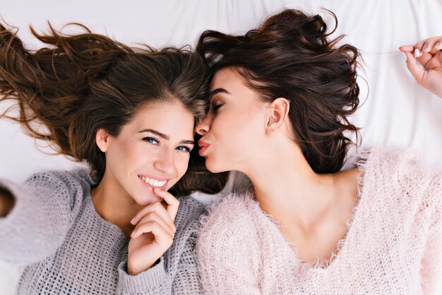 Omhoog selfie portret van twee vrolijke aantrekkelijke meisjes in wollen truien ontspannen op bed. 's Ochtends chillen, samen plezier maken, zorgen, mooie momenten van beste vrienden