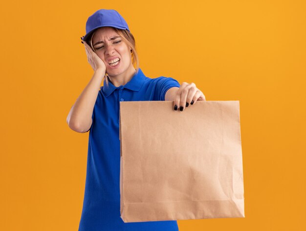 Onaangenaam jong mooi bezorgmeisje in uniform legt hand op gezicht en houdt papieren pakket op oranje