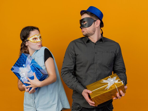 Onaangenaam jong partijpaar bekijkt elkaar die giftdozen houden die maskeradeoogmasker dragen dat op oranje wordt geïsoleerd