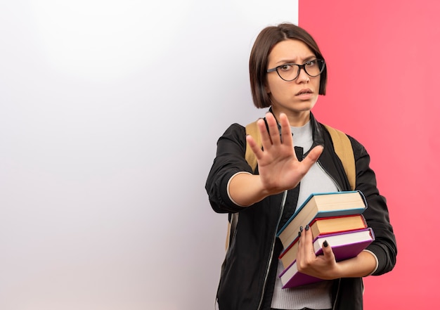 Onaangenaam jong studentenmeisje die glazen en achterzak dragen die boeken houden die zich voor witte muur bevinden gebaren niet geïsoleerd op roze achtergrond met exemplaarruimte