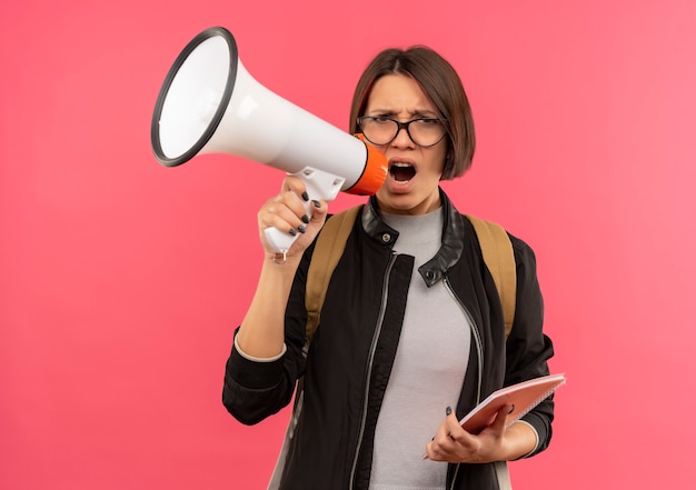 Gratis foto onaangenaam jong studentenmeisje die glazen en achterzak houden die notitieblok spreken door spreker die op roze achtergrond wordt geïsoleerd