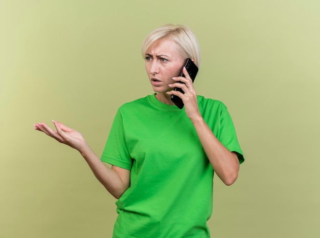 Gratis foto onaangename blonde slavische vrouw van middelbare leeftijd die aan de telefoon spreekt en naar kant kijkt die lege hand toont die op olijfgroene muur met exemplaarruimte wordt geïsoleerd