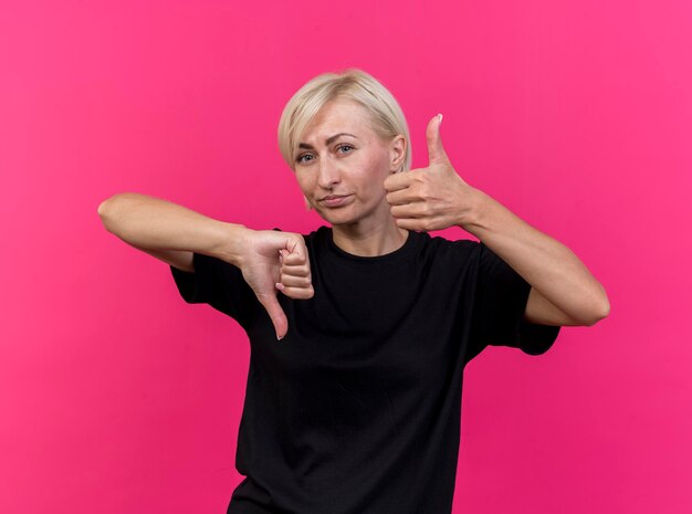Onaangename blonde Slavische vrouw van middelbare leeftijd die voorzijde bekijkt die duimen op en neer toont die op roze muur wordt geïsoleerd