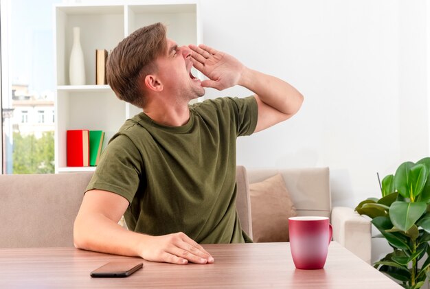 Onaangename jonge blonde knappe man zit aan tafel met kop en telefoon met hand dicht bij de mond en kijkt naar de zijkant en doet alsof hij iemand belt in de woonkamer